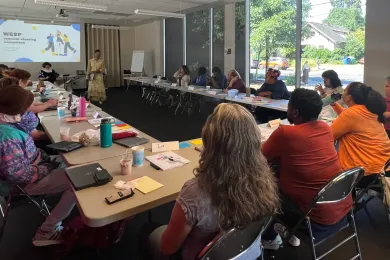 A group of people sit in chairs outside tables arranged in a U-shape. They are all facing the same direction toward a single person speaking to the group with a projection behind of a slide that says "WESP Renewal Steering Committee."