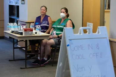 Two County staff members in brightly colored safety vests sit behind a table; water bottles, masks and other supplies are on the table; in the foreground is a sandwich board sign with the words "Come in and cool off"; a Spanish translation is below