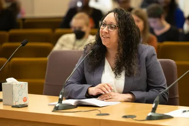 Rachael Banks sits at a table addressing an audience off camera. She sits behind a microphone on the table with a notebook placed in front of her.
