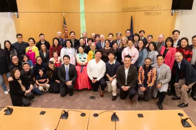 A portrait in the Multnomah County Board Room featuring local leaders and members of the Korean American community smiling at the camera