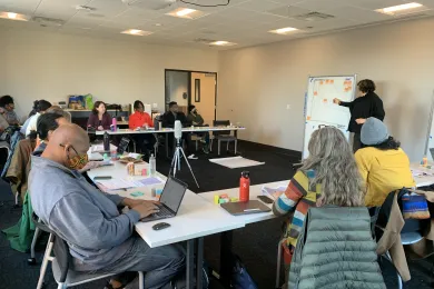 A group of people are seated behind tables set up in a U-shape. They are oriented toward a speaker at the front of the room who is pointing to a white board. On the board is a graph and several orange sticky notes.
