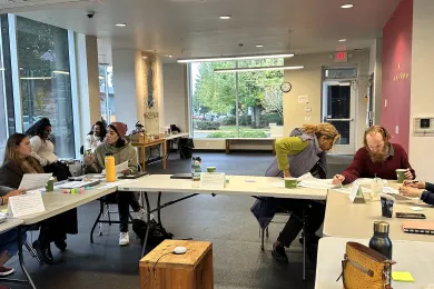 Two groups or three people each are seated around tables setup in a U-shape; two more people are in the background seated and speaking to each other.