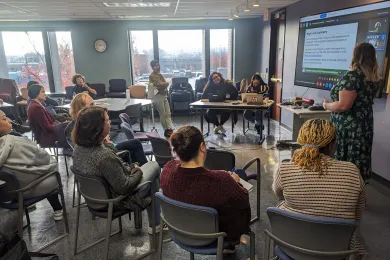 A group of people seated in chairs, along with one person standing, watch a presenter. The presenter is standing and is faced toward a screen on the wall.