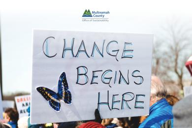 Image description: A sign with a drawing of a butterfly that says “Change begins here,” held by a gloved hand