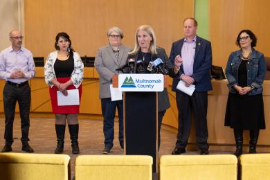 Five leaders standing side by side behind a podium as Commissioner Julia Brim-Edwards speaks about the purchase of a future sobering center.