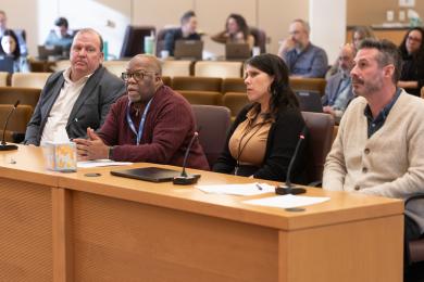 Four individuals seated at a panel in the Board Room, each with microphones in front of them. The panelists are engaged in discussion, with additional attendees in the background working on laptops and observing.