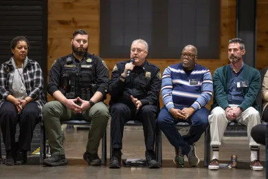 Five people seated in a row on chairs, facing the camera. One person is speaking into a microphone.