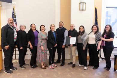 A group of ten people, appearing to be a mix of genders and ages, are standing indoors for a photo. They are positioned in a line facing the camera, with two flags visible in the background: the American flag on the left and the Oregon flag on the right. The setting is the Multnomah County Board Room, with light-colored walls and wood-paneled sections. The lighting is bright, likely from overhead fixtures and natural light from a window partially vi