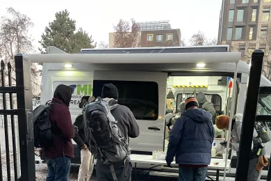 People stand in front of a folding table setup outside a white van with its doors open.