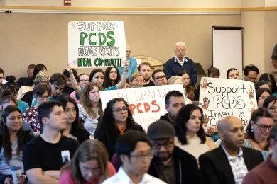 community members packed into an event room, some holding signs up 