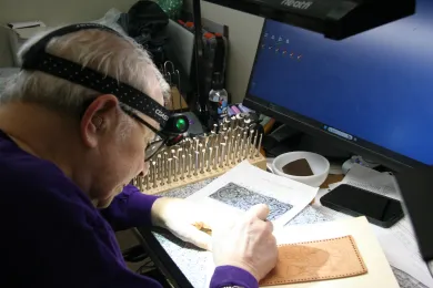 A close-up, shot photo of a person with white hair, wearing a purple long-sleeved shirt and a black headlamp with a green light, intently focused on engraving a design onto a rectangular piece of leather