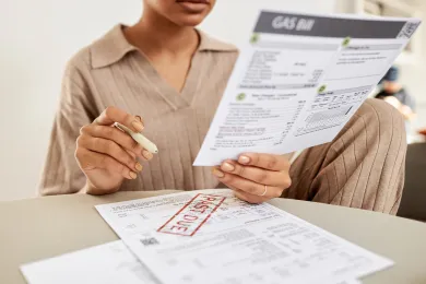 A person sits at a table covered in bills, and is holding a writing utensil and a utility bill.