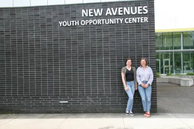 Two people stand side-by-side outdoors, smiling in front of a dark brick wall. Above them, white letters on the building read, "NEW AVENUES YOUTH OPPORTUNITY CENTER."