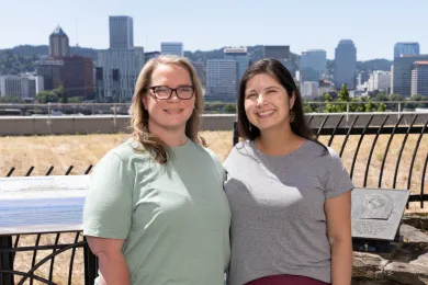 Two women stand on a high rise roof deck, shoulder to shoulder, with the landscape of Portland, OR downtown skyline in the background.