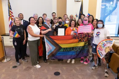 The Board of County Commissioners stands with a group of people holding up a pride flag.