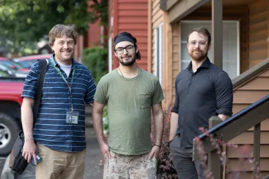 Three men stand side-by-side outdoors, smiling at the camera.