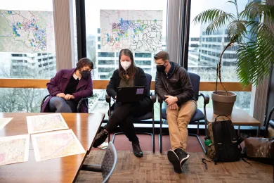 Three people sit in chairs next to each other, all looking at one laptop.