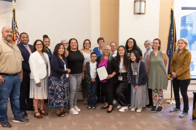 A smiling group of people pose for the camera between an American flag and a State of Oregon flag. A person toward the center of the group holds up the proclamation.