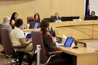 An eye-level shot of a formal public or board meeting in a room with light wood paneling. In the background, four officials are seated behind a long, curved wooden dais. In the foreground, several attendees are seen from behind, seated at a table with open laptops and tablets, facing the officials.