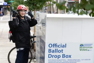  A person wearing a black jacket, dark pants, sneakers, and a red and white polka-dot helmet stands next to a white and blue official Ballot Drop Box on a sidewalk.