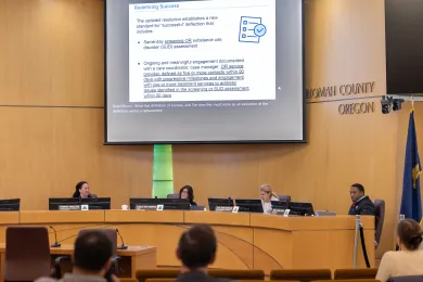 A Multnomah County Board room during a presentation on "Redefining Success" for the HB 4002 Deflection Program. A large screen displays new standards for successful deflection, including same-day screenings and 90-day engagement milestones. Four commissioners sit at the dais facing the audience.
