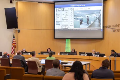 A wide shot of the Multnomah County Board of Commissioners meeting room. Five commissioners sit at a curved dais beneath a large projector screen showing "Sobering Care" facility floor plans and 3D renderings. Several people in the audience face the dais.