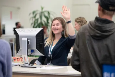 A Multnomah County Elections worker smiles and raises her hand to assist a member of the public at a service desk.