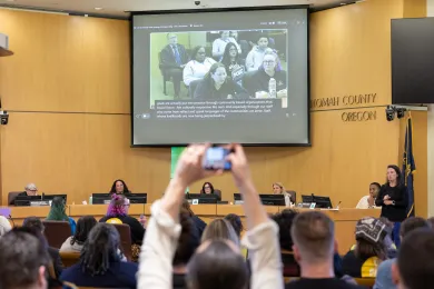 An audience member takes a photo during a Multnomah County public budget hearing where a witness testifies via video screen.