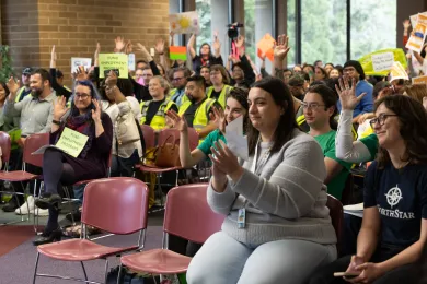 Community members sitting clapping and waving hands in support of others testifying in room. 