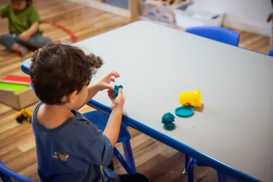 A young child with curly brown hair sits at a table, focused on shaping a small piece of teal playdough in their hands.