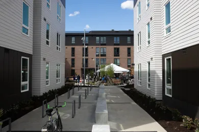 A concrete courtyard with bike racks sits between two modern, multi-story apartment buildings, leading toward an outdoor community gathering with a white canopy tent in the background.