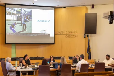 La Junta de Comisionados del Condado de Multnomah y representantes de la División de Educación Preescolar y Aprendizaje Temprano se sientan alrededor de una gran mesa redonda dentro de la sala de juntas del Condado de Multnomah. 