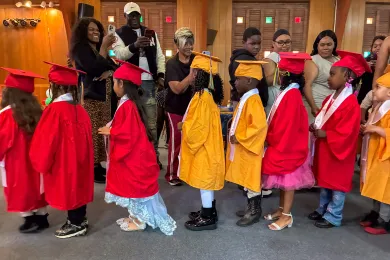 Varios niños de preescolar, vestidos con togas y birretes de graduación rojos o amarillos, están formados en fila india. 