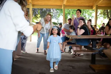 A Preschool for All student in a blue dress and tiara walks through a graduation celebration as families and teachers look on.