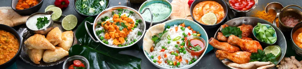 A variety of different foods on a table, including rice topped with vegetables, samosas, curries, and chicken drumsticks