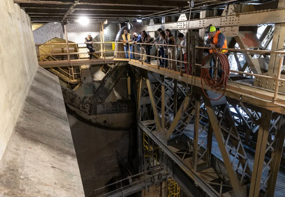 A group of people in safety vests standing on a catwalk under a bridge, looking down into a large open space with a giant concrete structure on one side.