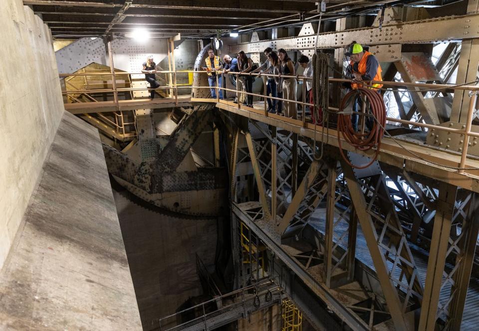A group of people in safety vests standing on a catwalk under a bridge, looking down into a large open space with a giant concrete structure on one side.