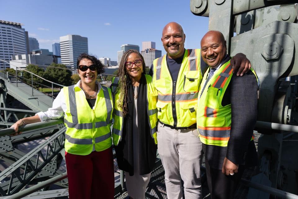 Four people in safety vests pose for the camera in an elevated location, with mechanical bridge elements and the city skyline in the background.