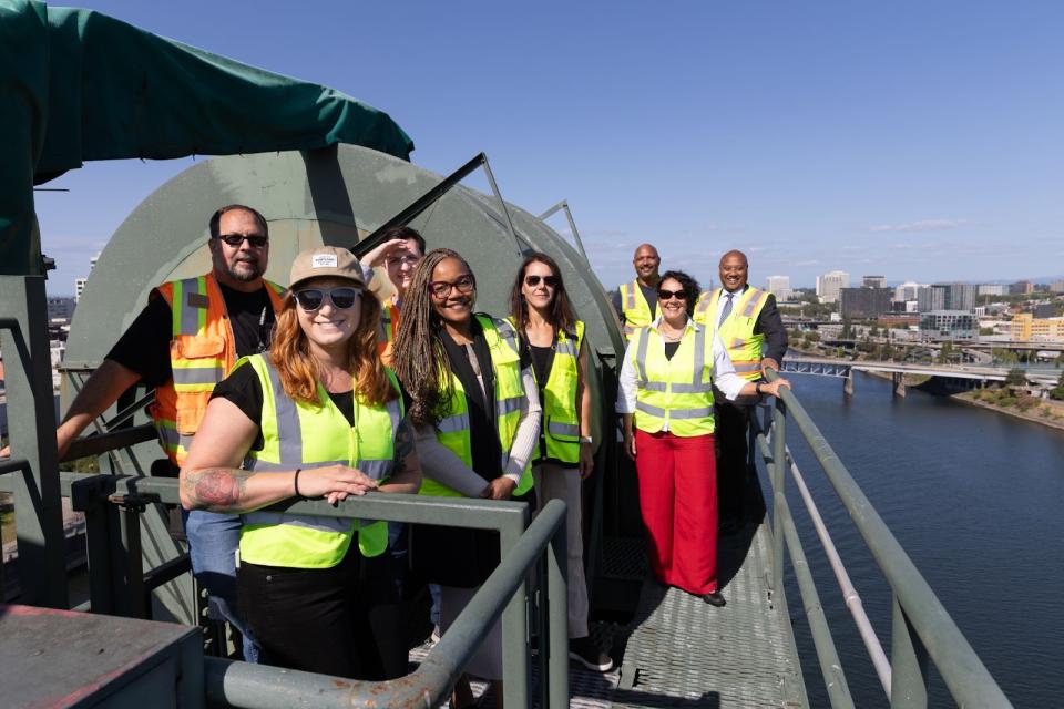 A group of eight people in safety vests posed for the camera on top of a bridge, with mechanical structures, the river and the city skyline in the background.