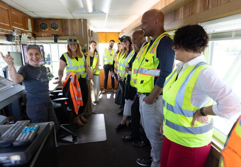 A group of people in safety vests gather in a bridge control room, giving their attention to a bridge operator who is pointing at something off-camera.