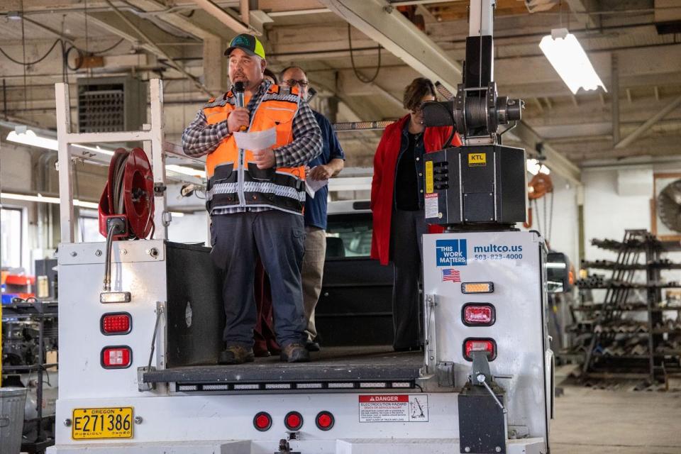 A man with a with a high-visibility vest and hat speaks from the back of a maintenance truck inside a large garage.