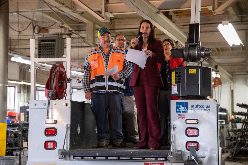 A woman with a microphone speaks from the back of a maintenance truck, flanked by a man with a high-visibility vest and hat. They are inside a large garage.