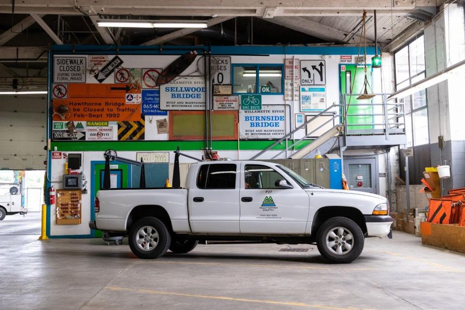 A white work truck with a Multnomah County logo parked in front of a two-story maintenance shed decorated with old traffic signs, inside a large garage.