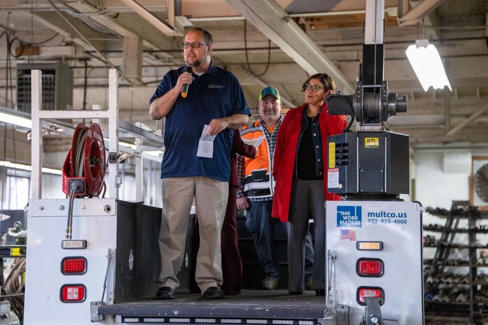 A man with a microphone speaks from the back of a maintenance truck inside a large garage.