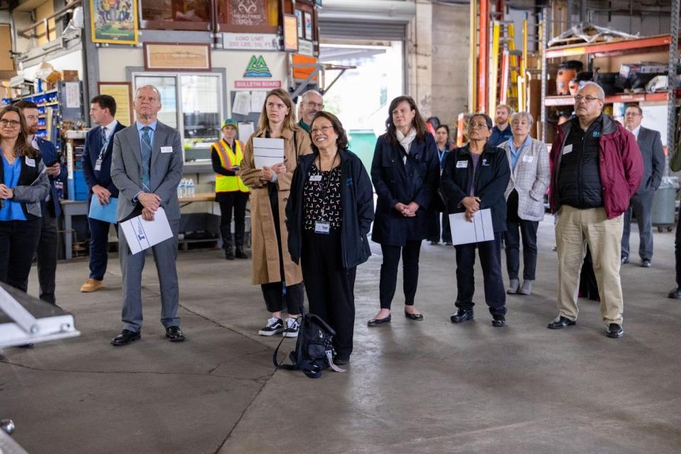 A group of people stand in a large garage, giving their attention to a speaker.