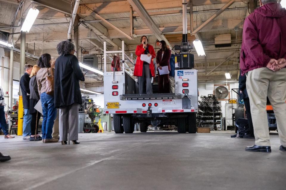 A woman with a microphone addresses a group of people from the back of a maintenance truck. They are inside a large garage.