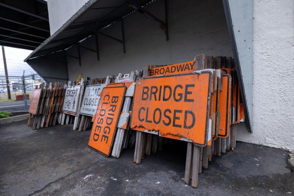 Several stacks of Bridge Closed or Road Closed signs, leaned up against a wall with the underside of a bridge visible above in the background.