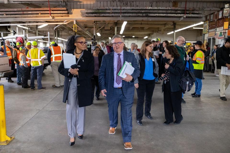 A man and a woman talk with each other as they walk away from a larger group, inside a large maintenance garage.