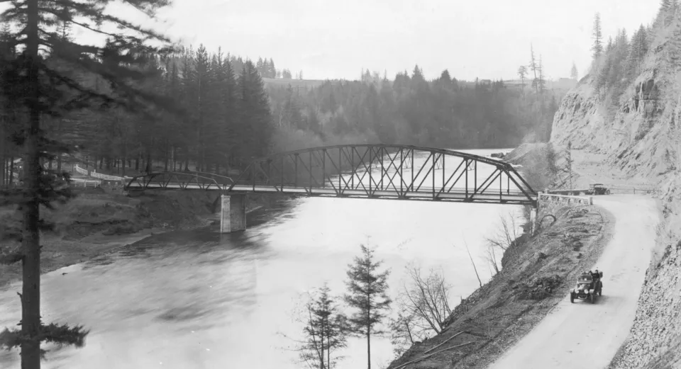 A black and white photo of the Stark St Bridge. The narrow and unfinished Stark Street and early automobiles indicate this is a very old photo.
