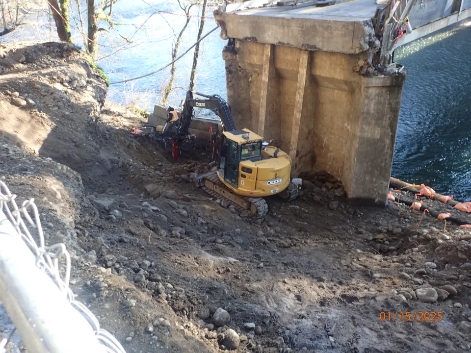 An excavator at work on an embankment next to a concrete support for the Stark Street Bridge.